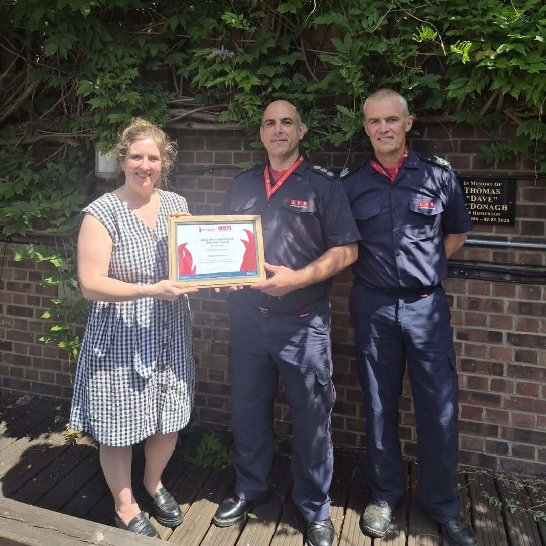 Three people stand outdoors in sunlight; two men in dark uniforms hold a framed certificate, while a woman in a checkered dress stands beside them. They are in front of a brick wall with vines and a commemorative plaque.