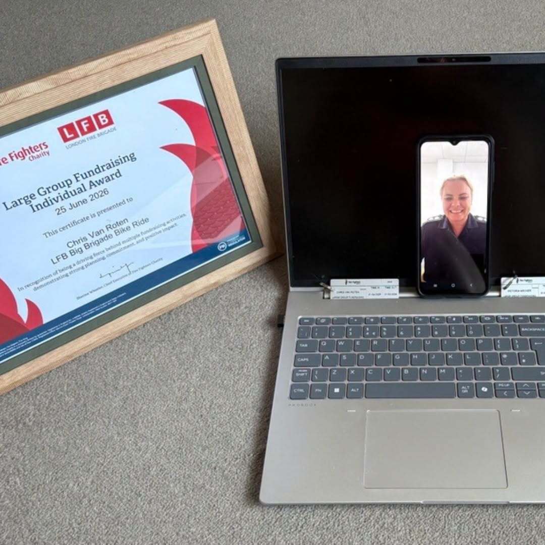 A framed award certificate lies on a carpet next to an open laptop. A mobile phone is propped on the laptop keyboard, displaying a smiling woman on a video call.