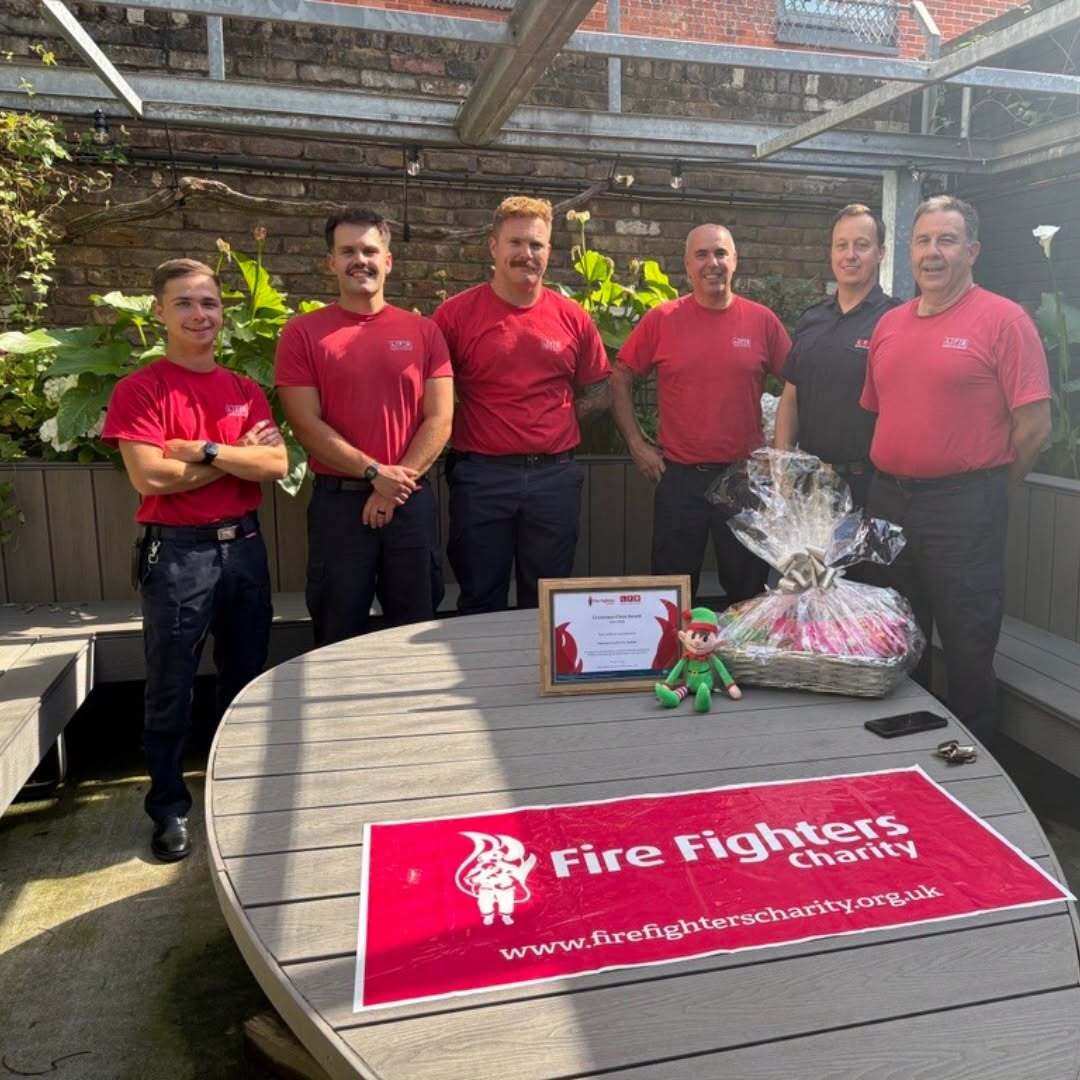 Six firefighters pose and smile behind a round table displaying a plaque, a gift basket, a plush toy, and a Fire Fighters Charity banner in an outdoor setting with brick walls and greenery.