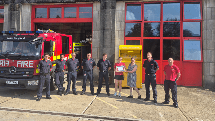 A group of firefighters and two civilians stand outside a fire station with a fire engine and a yellow box in the background. One civilian holds a certificate, and everyone is smiling at the camera.