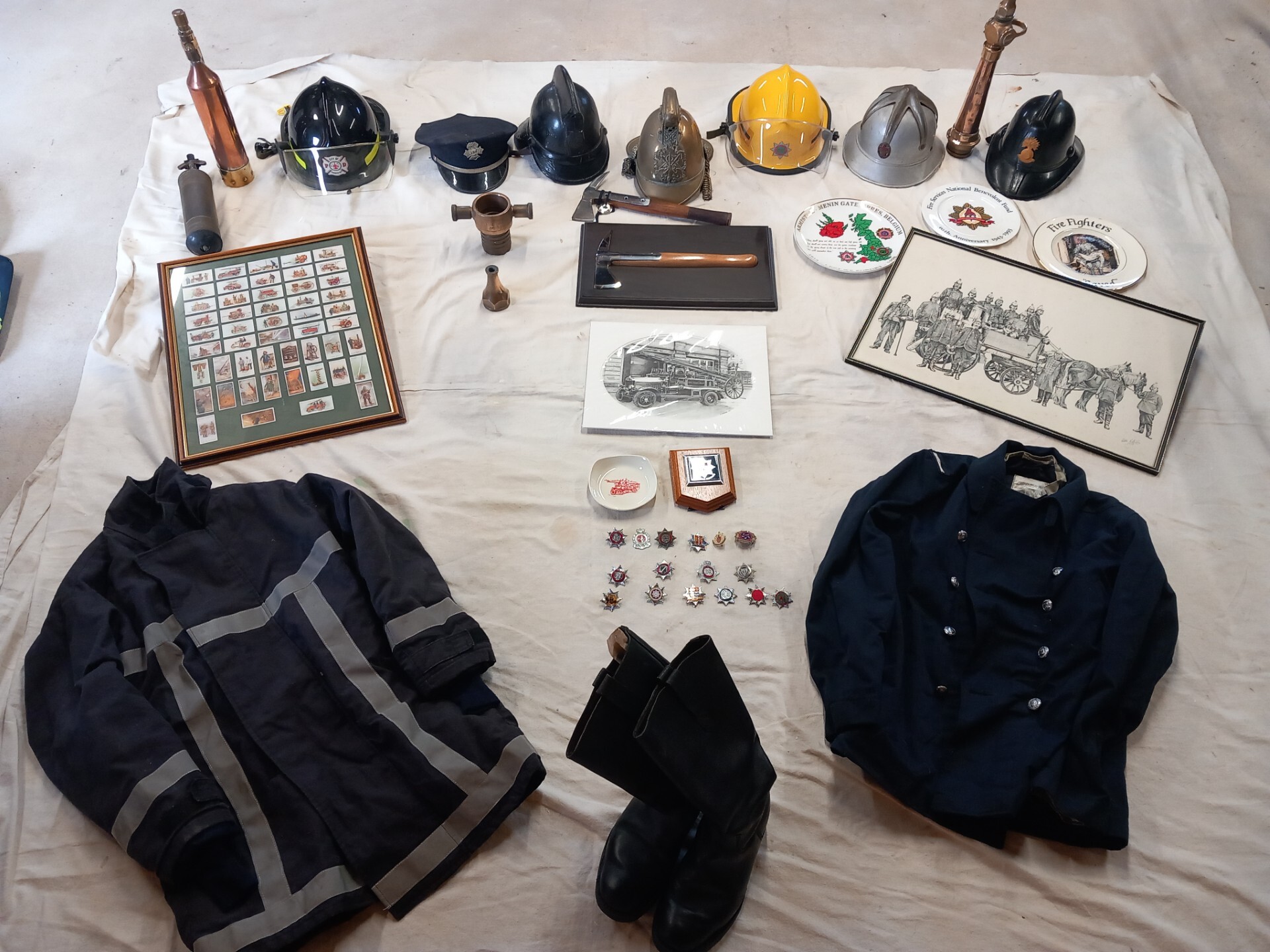 A collection of vintage firefighter memorabilia is arranged on a white cloth, including helmets, uniforms, boots, medals, framed badges, plates, and antique tools.