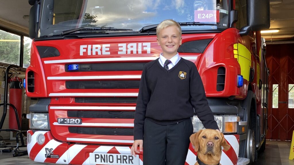 A smiling boy in a black school uniform stands in front of a red fire truck, with one hand resting on a golden Labrador dog sitting beside him.