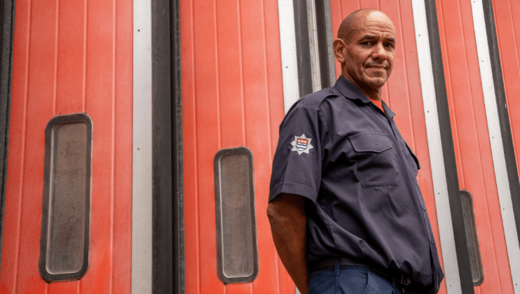 Merrick a firefighter in uniform stands in front of tall, red fire station doors, looking off to the side with a serious expression.