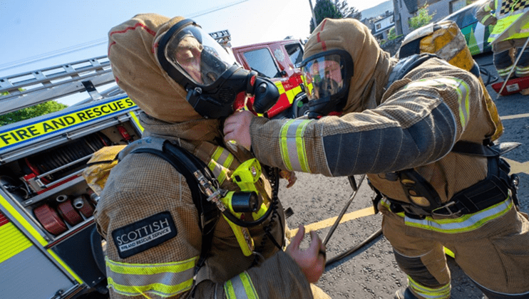 Two firefighters in full gear and breathing apparatus prepare for action near a fire engine. One firefighter assists the other with their mask. Both are outside on a sunny day with emergency vehicles nearby.