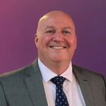 A bald man in a grey suit, white shirt, and navy polka dot tie is smiling in front of a purple background.