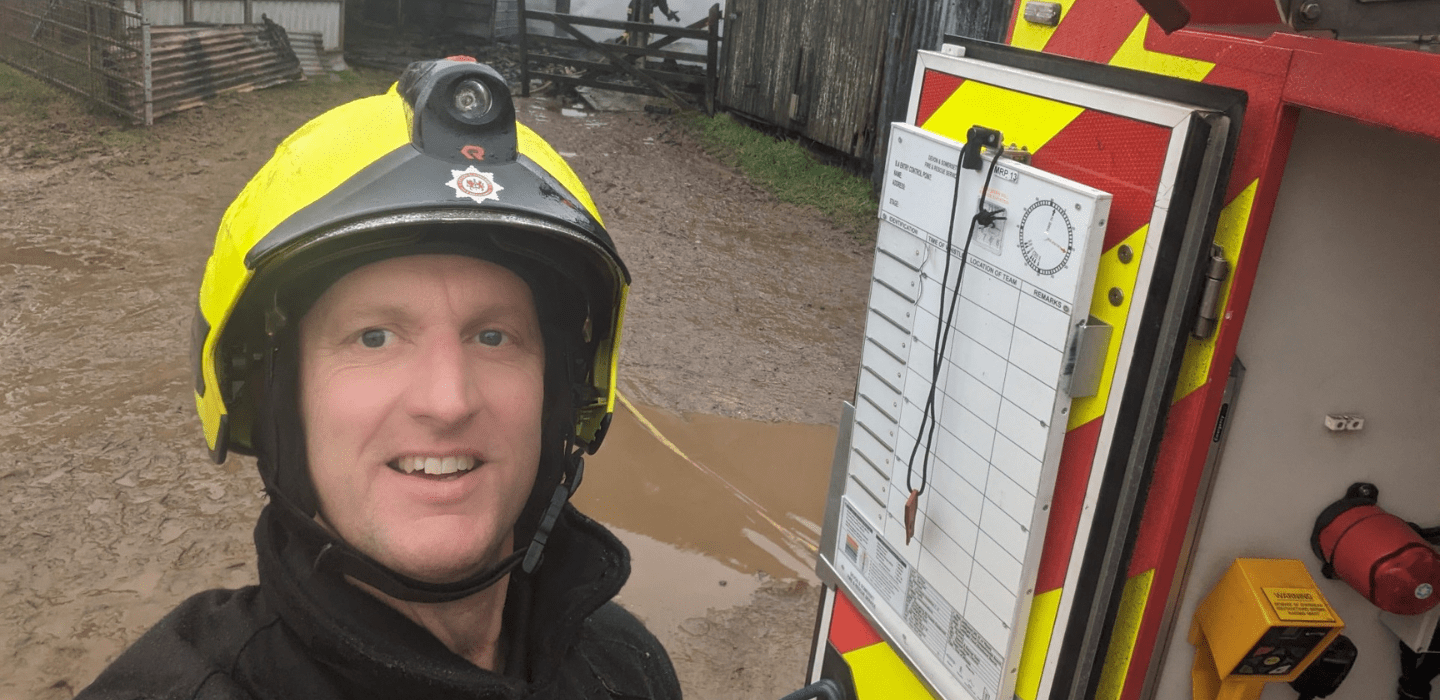 A firefighter in a yellow helmet smiles for a selfie outdoors near a muddy area, standing beside an incident command board attached to a fire vehicle. 