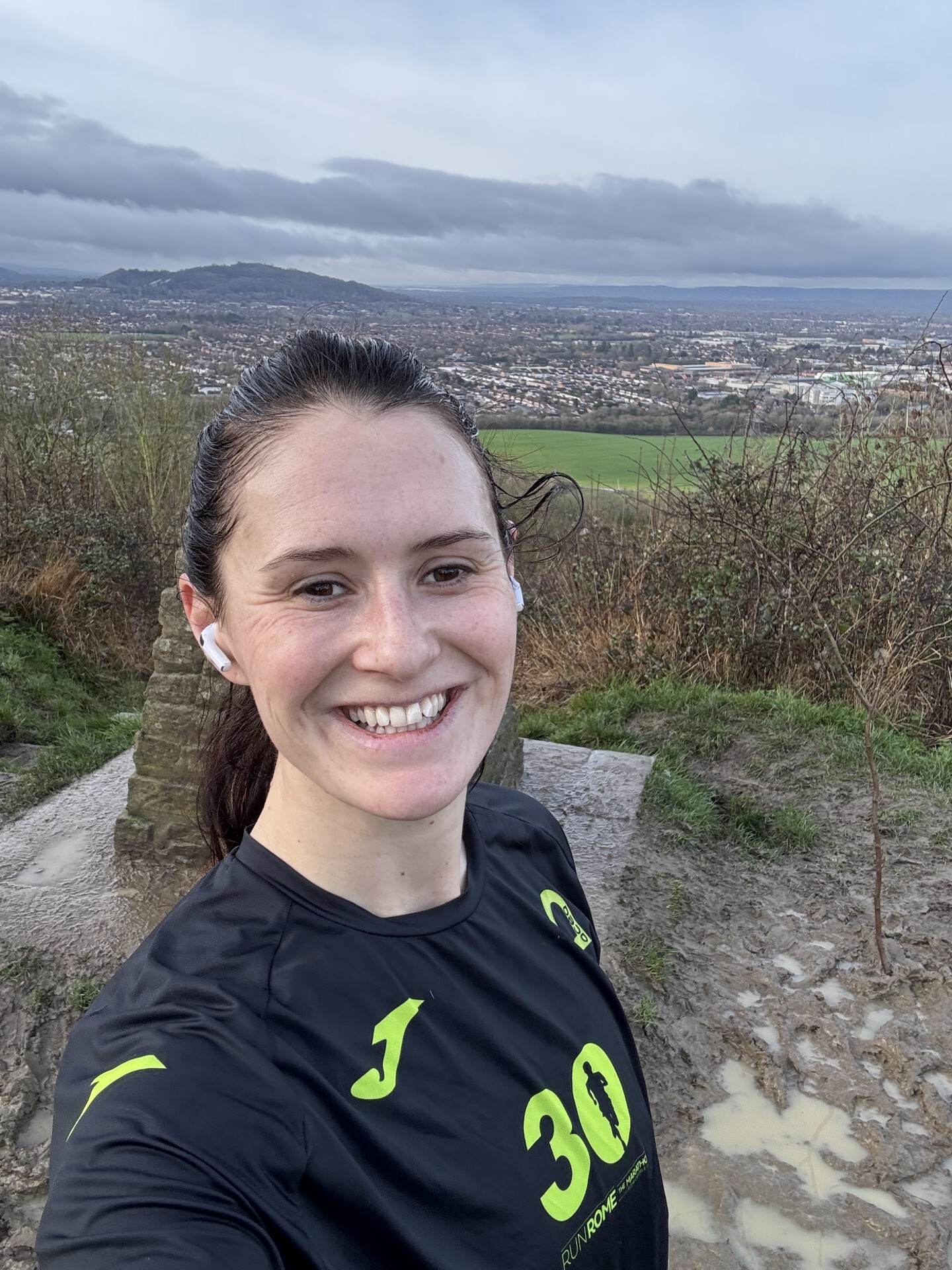 A woman in a black athletic shirt smiles for a selfie on a muddy hilltop with earbuds in. The background shows a panoramic view of a town, fields, and a cloudy sky.