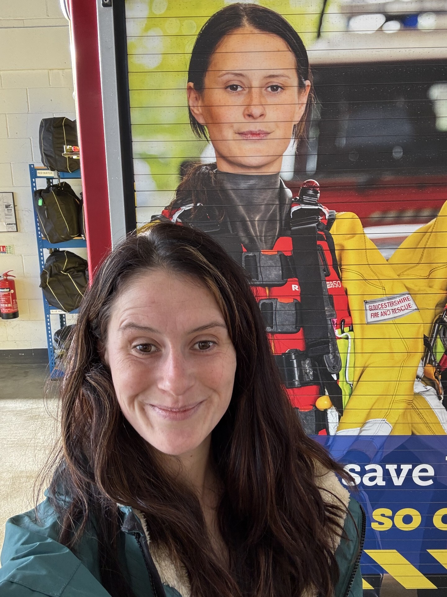 A woman with long brown hair smiles, taking a selfie in front of a large poster showing herself in a fire and rescue uniform at an indoor facility. A fire extinguisher and shelves with bags are visible in the background.