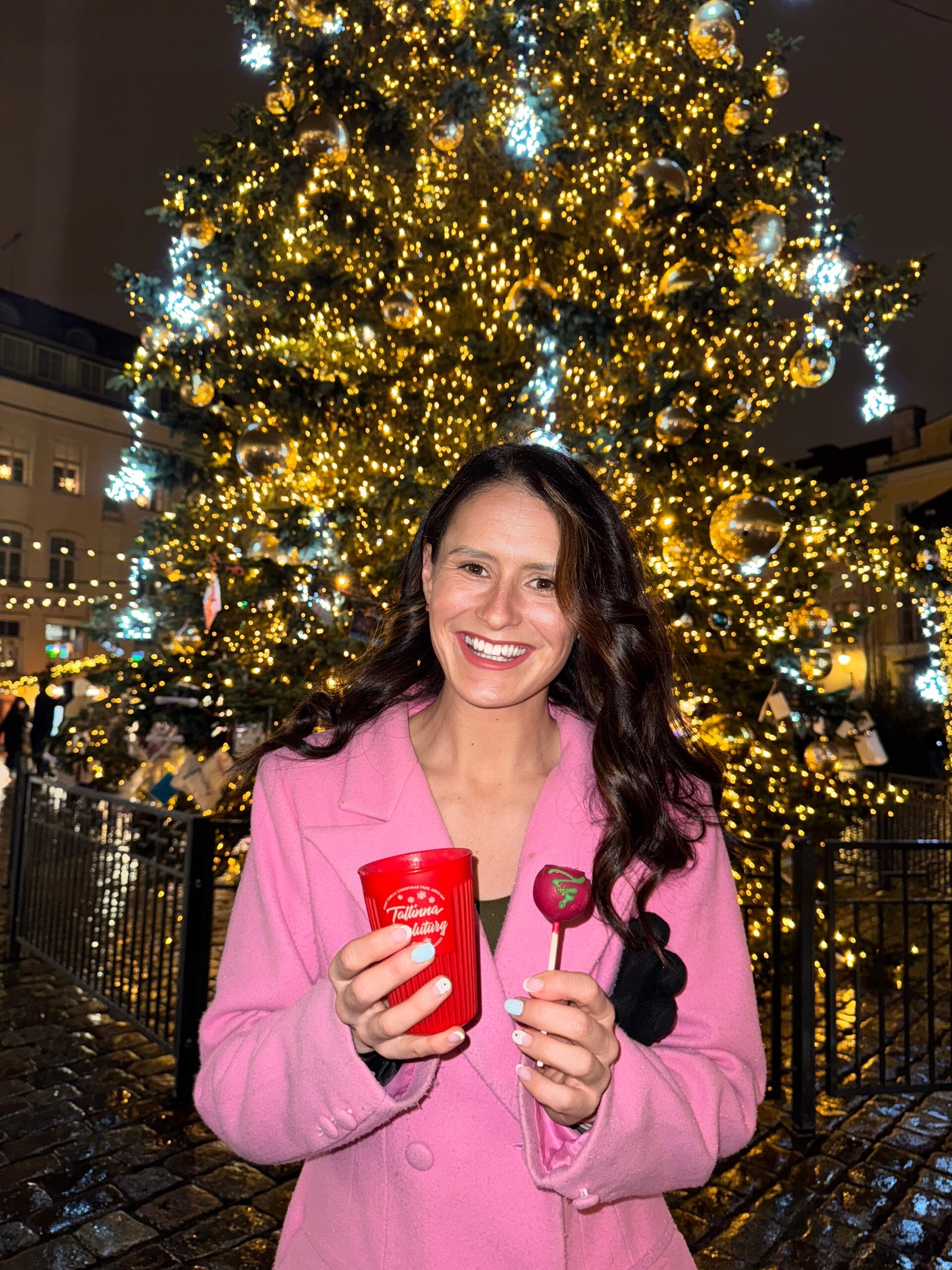A smiling woman in a pink coat holds a red cup and a heart-shaped treat, standing in front of a large, decorated Christmas tree with glowing lights at night.