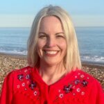A woman with light blonde hair, wearing a bright red sweater with floral embroidery, smiles while standing on a pebbled beach with calm blue sea and clear sky in the background.