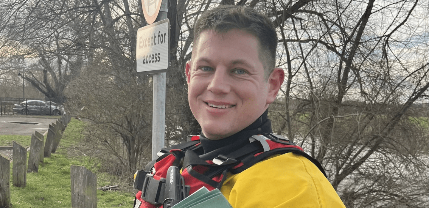 A person wearing a yellow and red rescue suit stands outdoors near a river, smiling at the camera. Behind them are bare trees, a parking area, and a sign that reads 