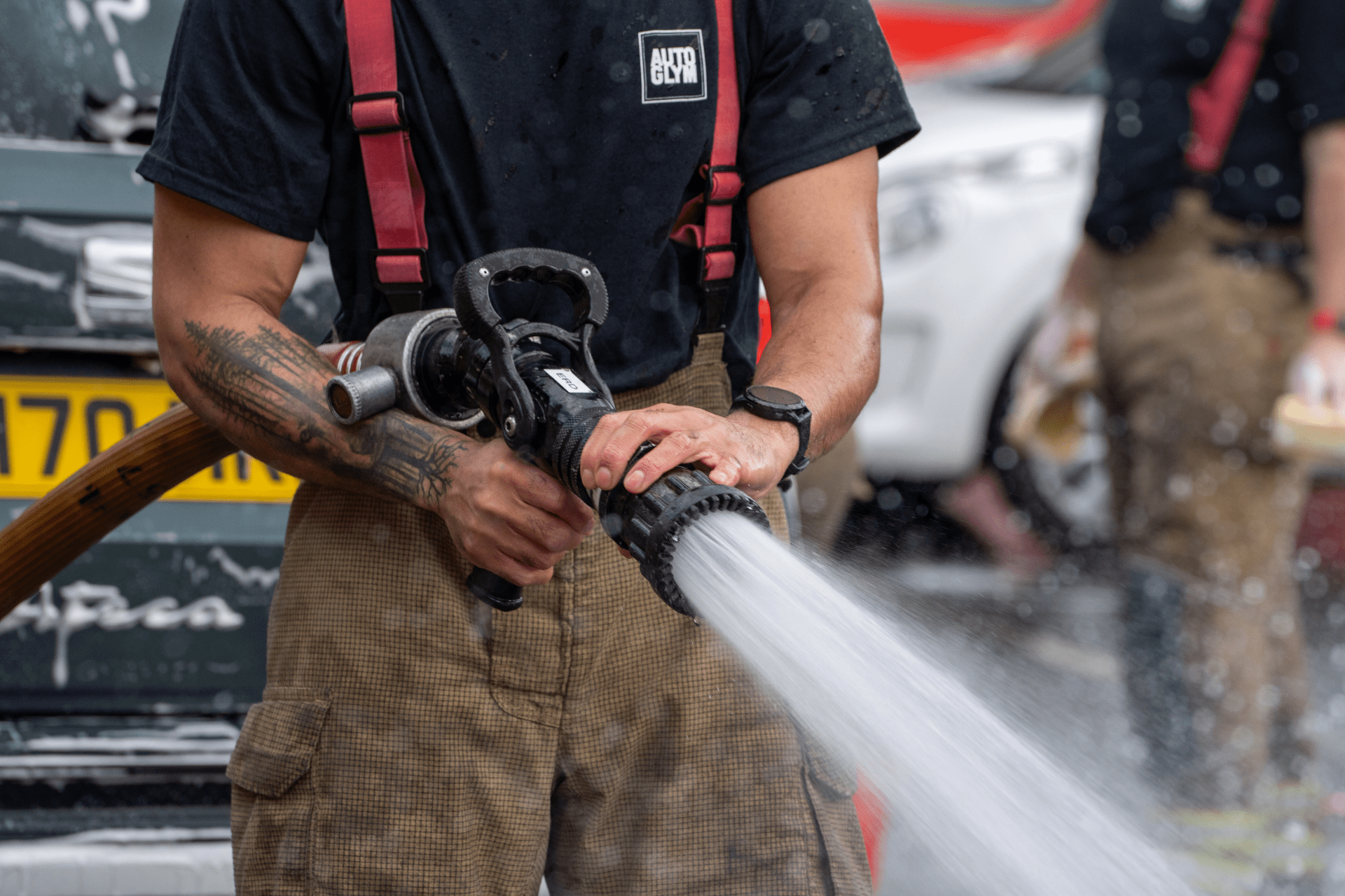 A firefighter wearing suspenders and brown trousers uses a hose to spray water, with water splashing around. Part of a fire truck and another firefighter are visible in the background.