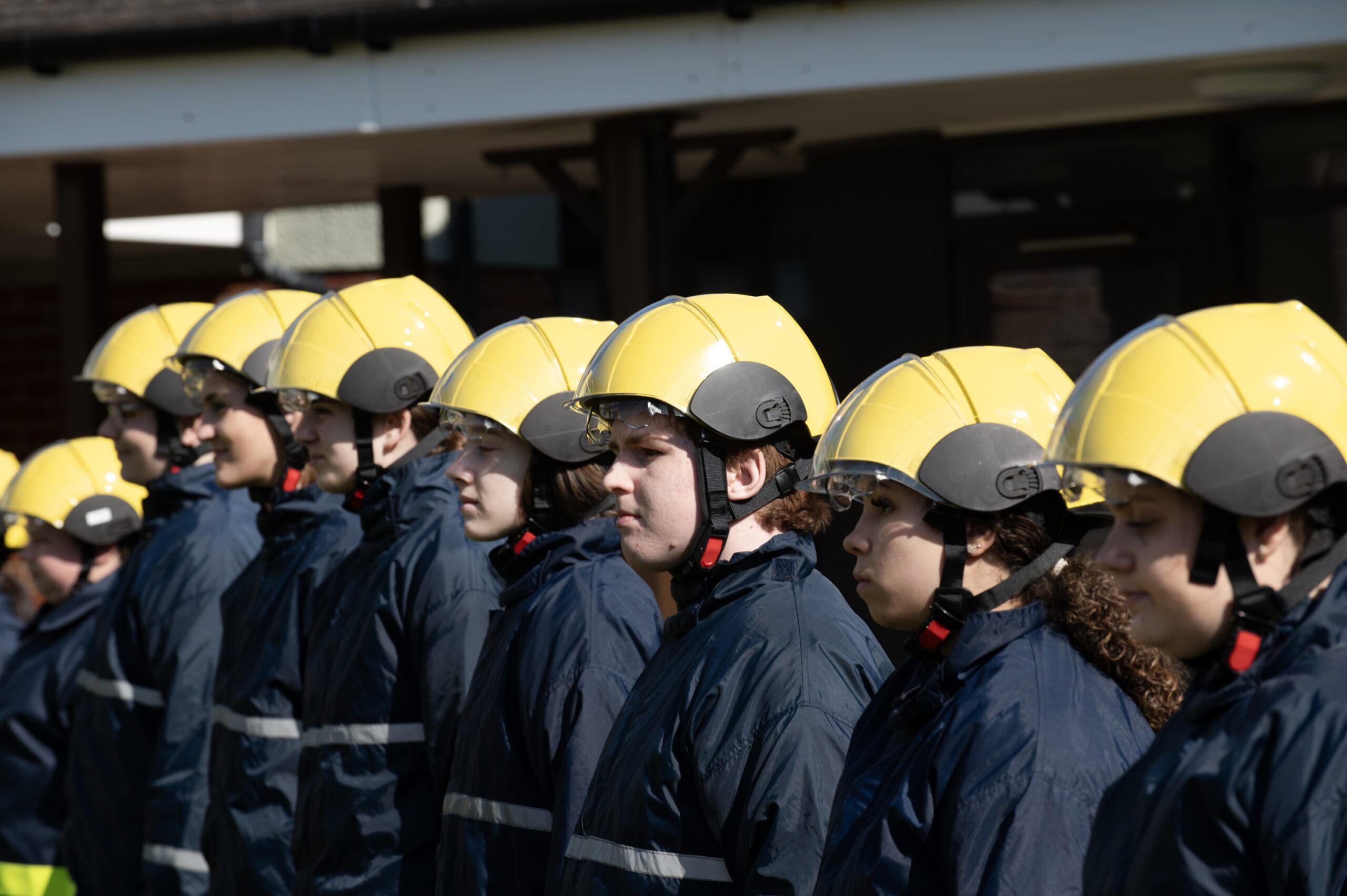 A row of firefighters in dark uniforms and yellow helmets stand in line outdoors, facing the same direction, with a building visible in the background.