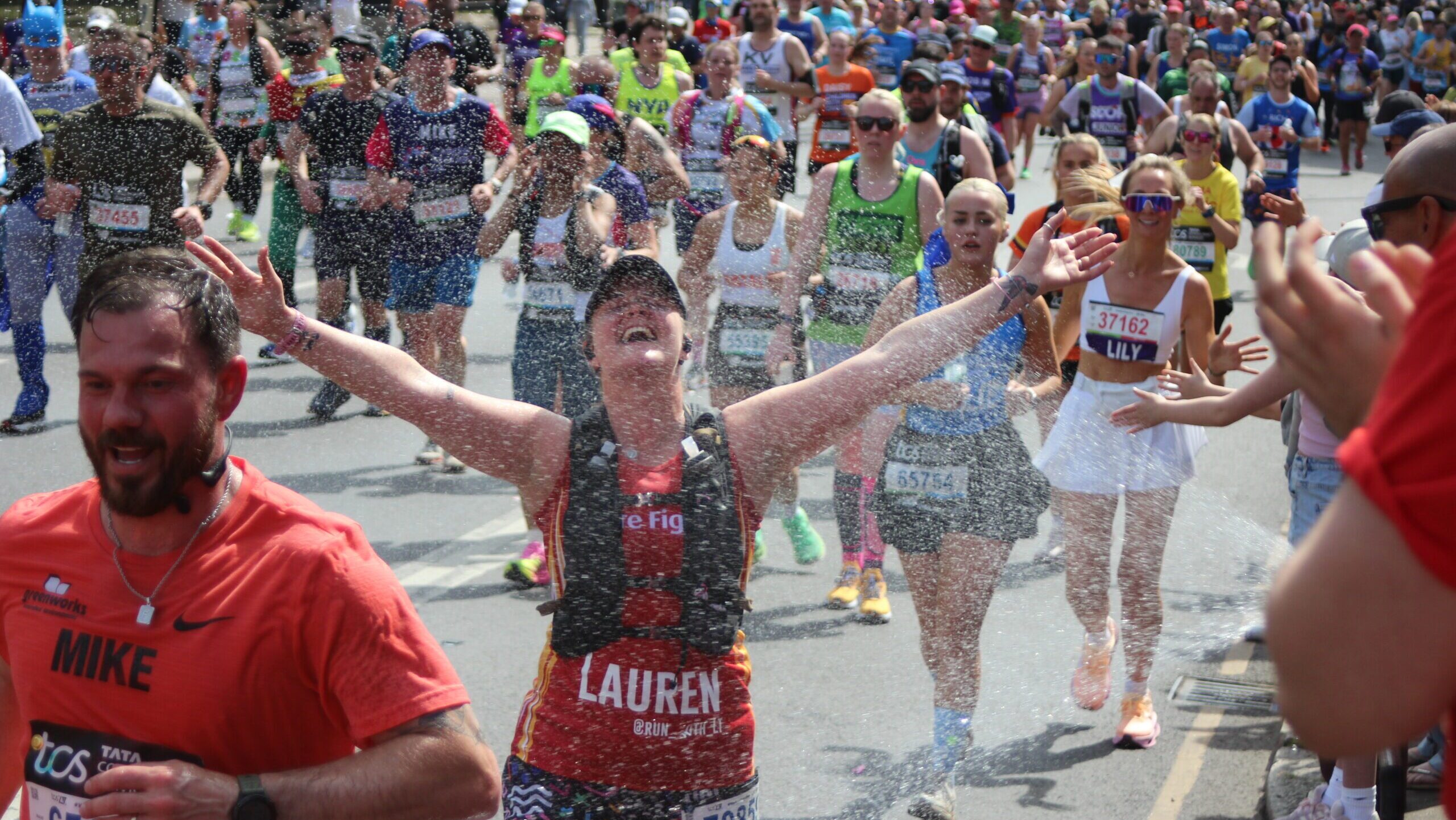 Runners in a marathon are splashed with water as they pass, part of a fire fighters charity event highlighted in the latest news. A woman in front, arms raised, smiles joyfully. Crowds of runners and some spectators are in the background.