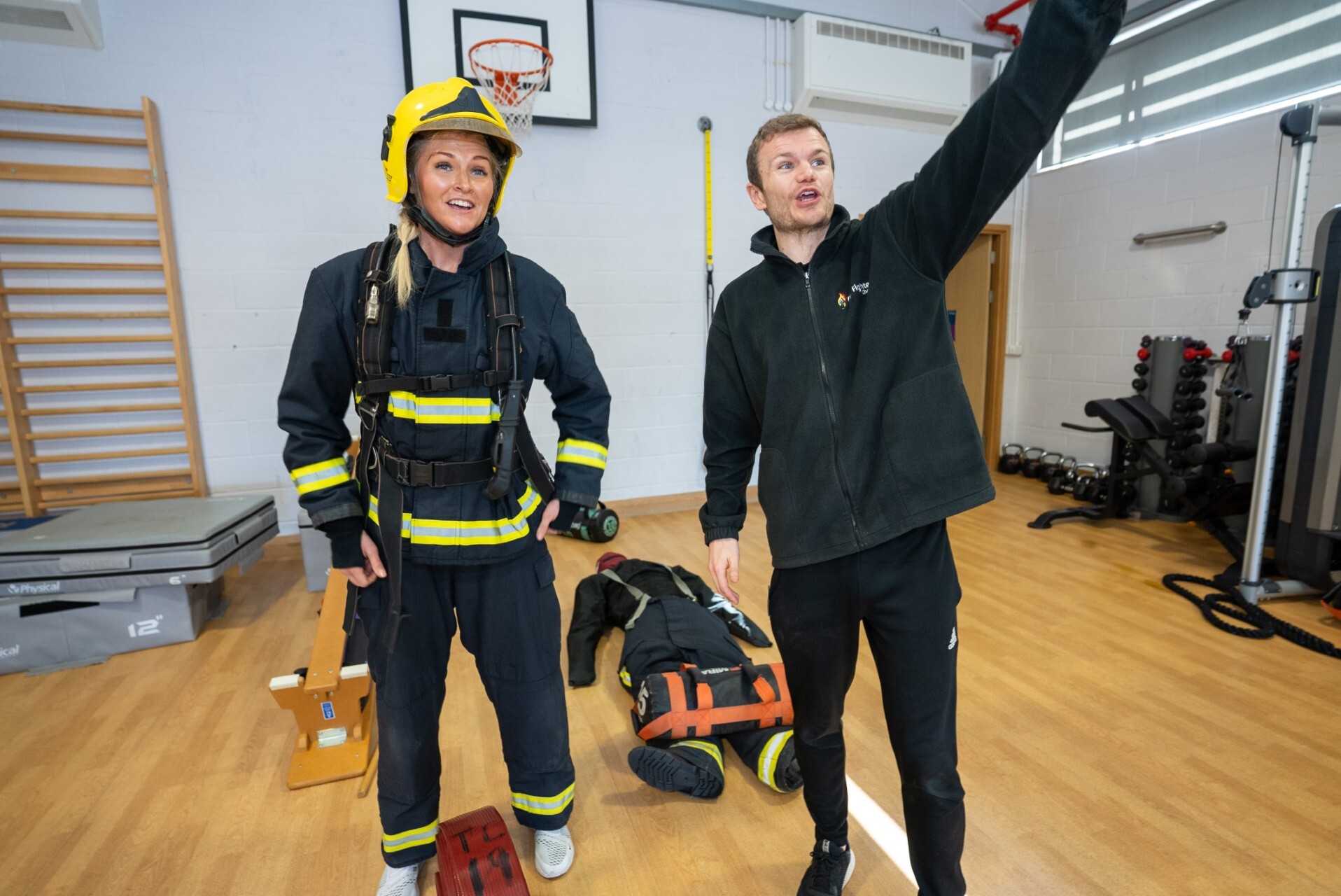 A firefighter in uniform stands next to a man pointing while another person lies on the floor behind them in a gym, suggesting a training or rescue drill scenario. 