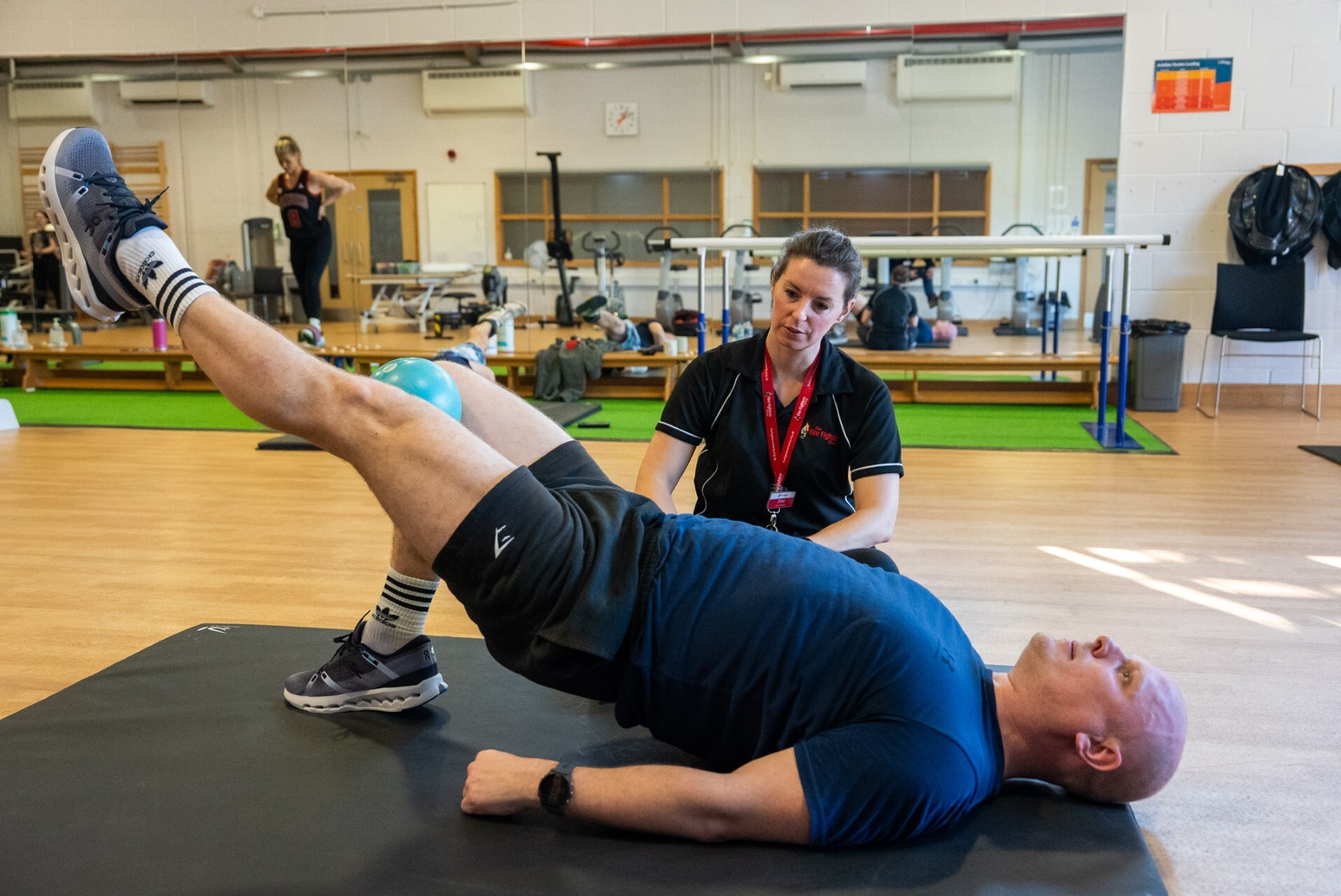 A man performs a glute bridge exercise on a mat while a trainer kneels beside him, observing and offering guidance in a gym with exercise equipment in the background.