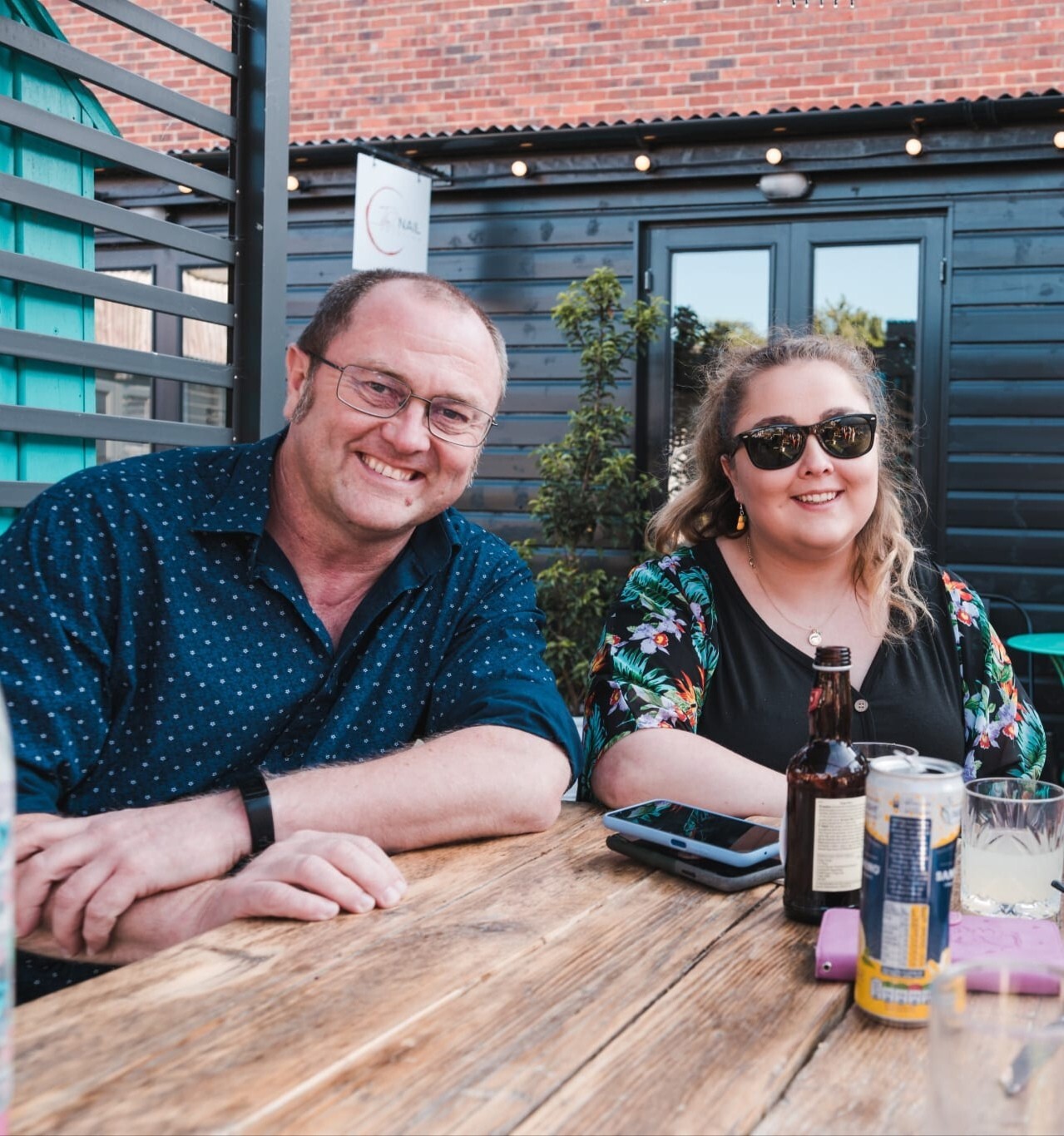 Two people sit at an outdoor wooden table, smiling. The man wears glasses and a blue shirt; the woman has sunglasses and a floral top. Drinks and a phone are on the table. A black building and plants are in the background.