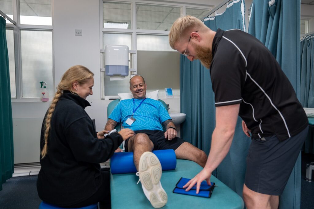 A man lies on a medical table smiling while two therapists, one male and one female, help him exercise his legs in a clinic with blue privacy curtains.