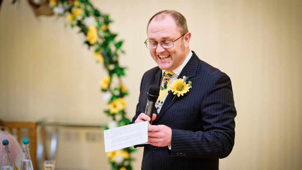 A man in a suit with a sunflower boutonnière smiles while speaking into a microphone and holding a paper, standing beside a floral arch decorated with yellow flowers.