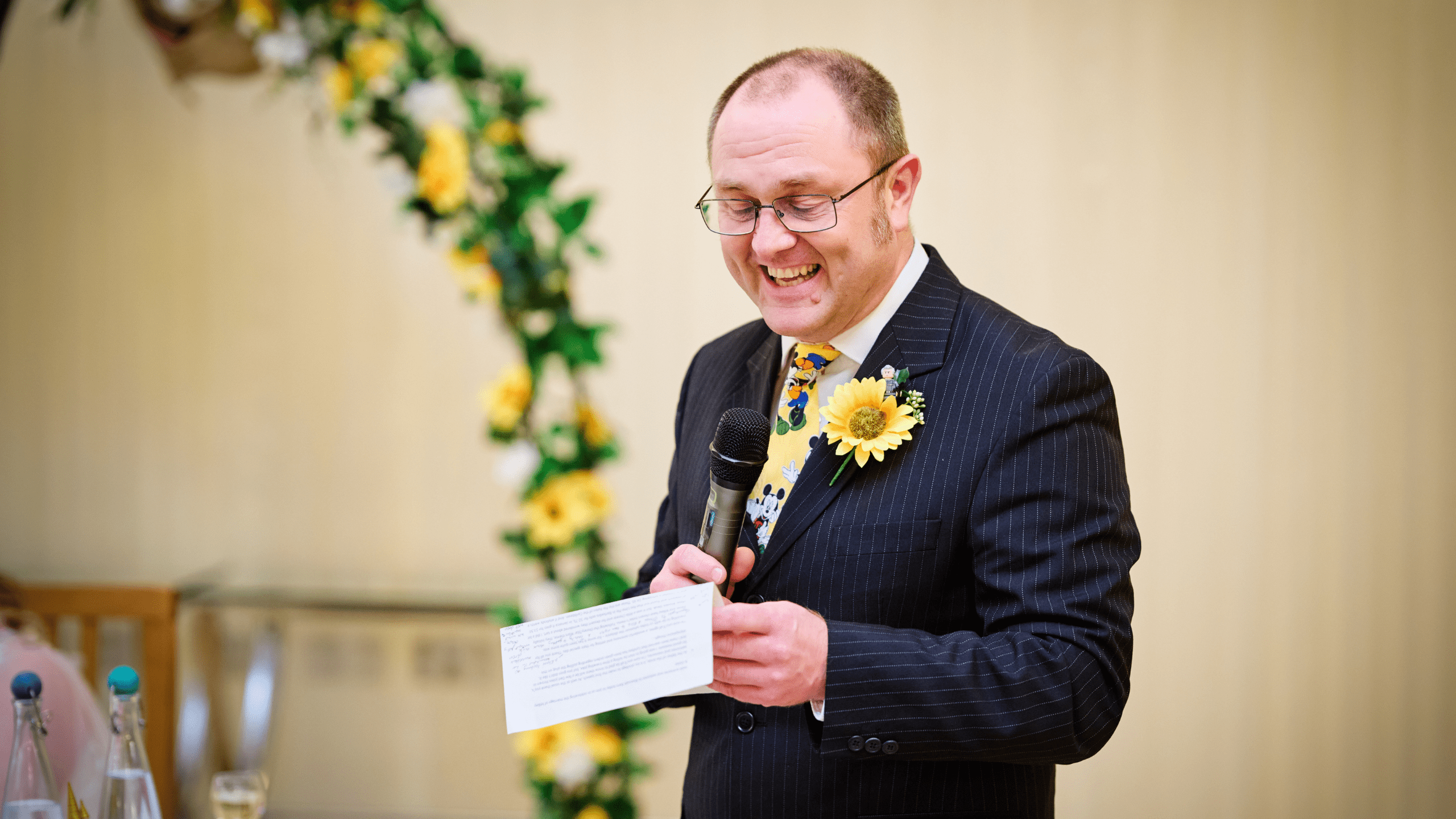 A man in a suit with a sunflower boutonnière smiles while speaking into a microphone and holding a paper, standing beside a floral arch decorated with yellow flowers.