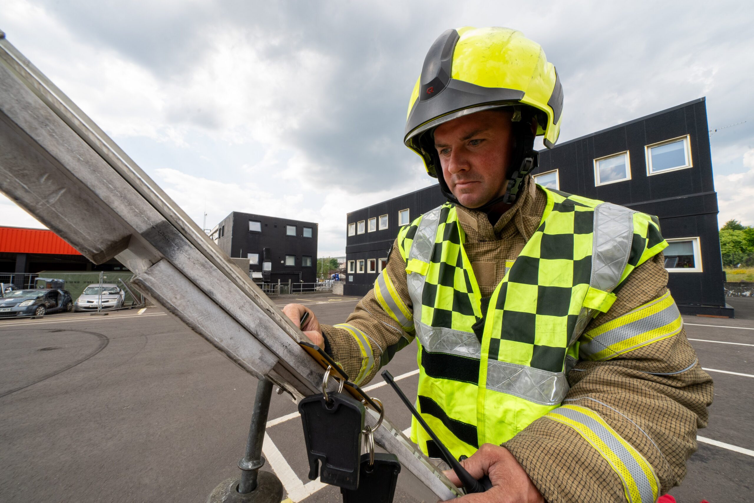 A firefighter wearing safety gear and a high-visibility vest adjusts equipment on a metal ladder in an outdoor training area, with modern buildings in the background. 