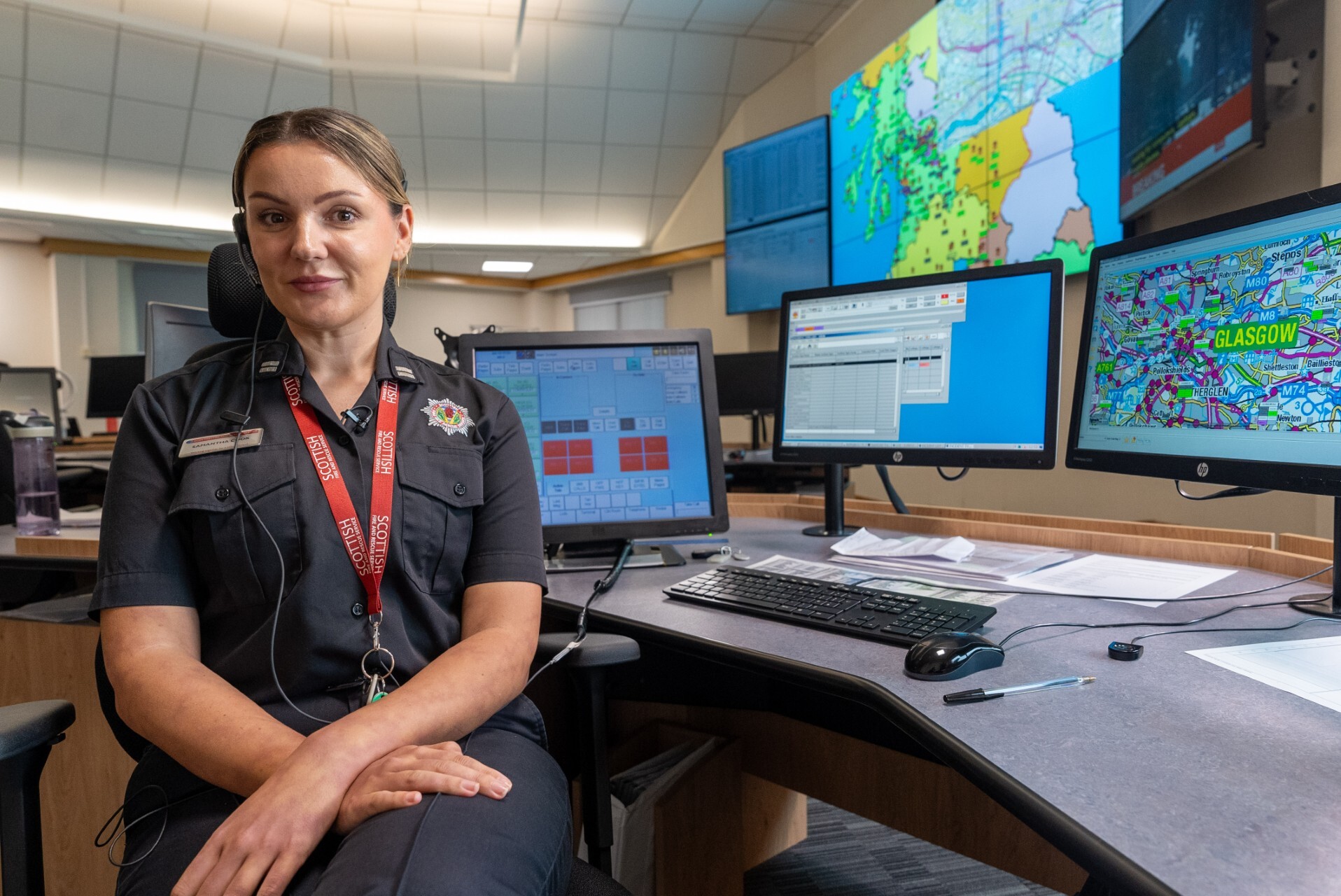 A woman in a dark uniform sits at a control desk with multiple monitors displaying maps and data in an emergency operations center.