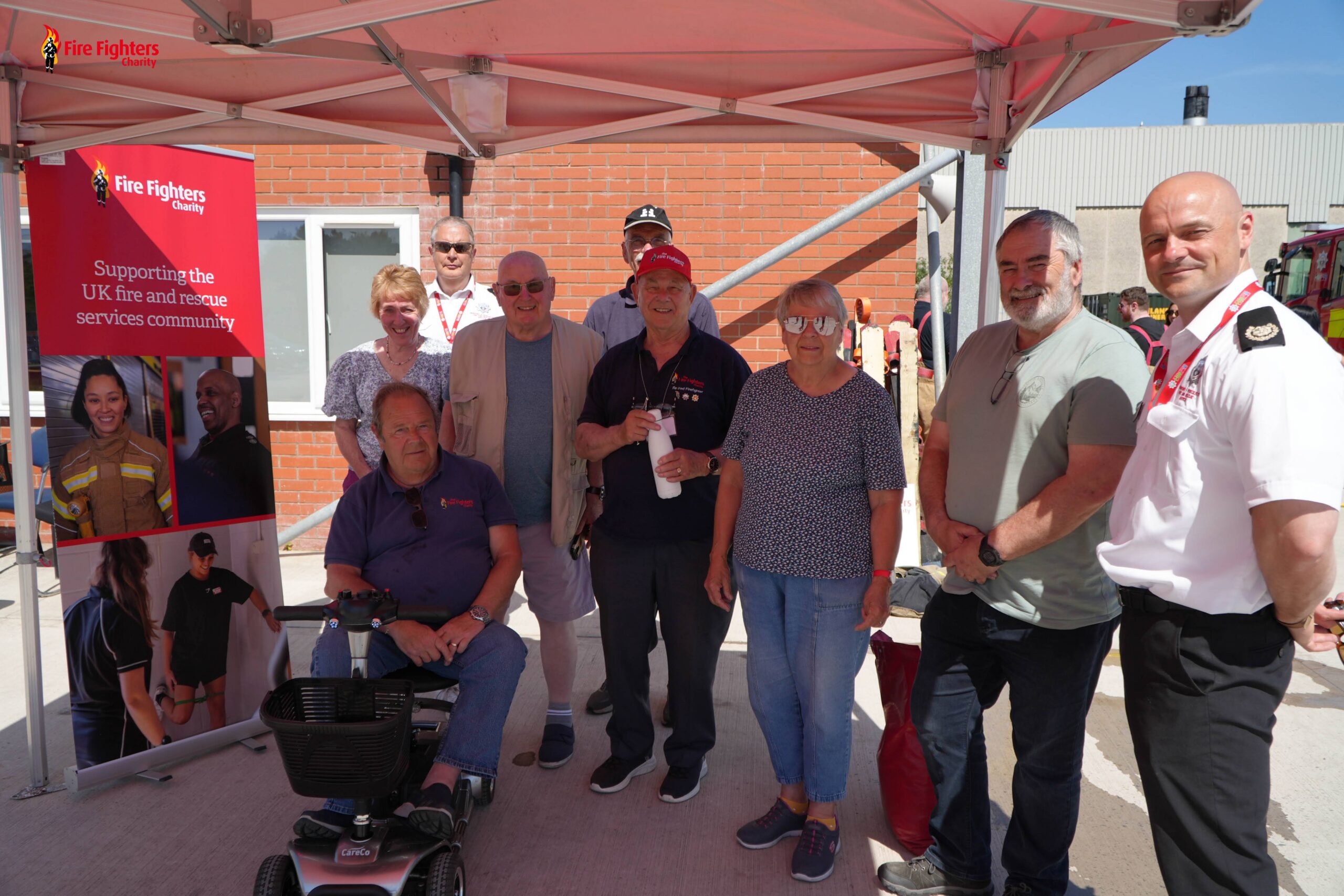 A group of people, including a man in a wheelchair, smiling and standing under a tent at a fire service event. A Fire Fighters Charity banner is displayed on the left, highlighting volunteer opportunities for those looking to support or volunteer for fire fighters charity.