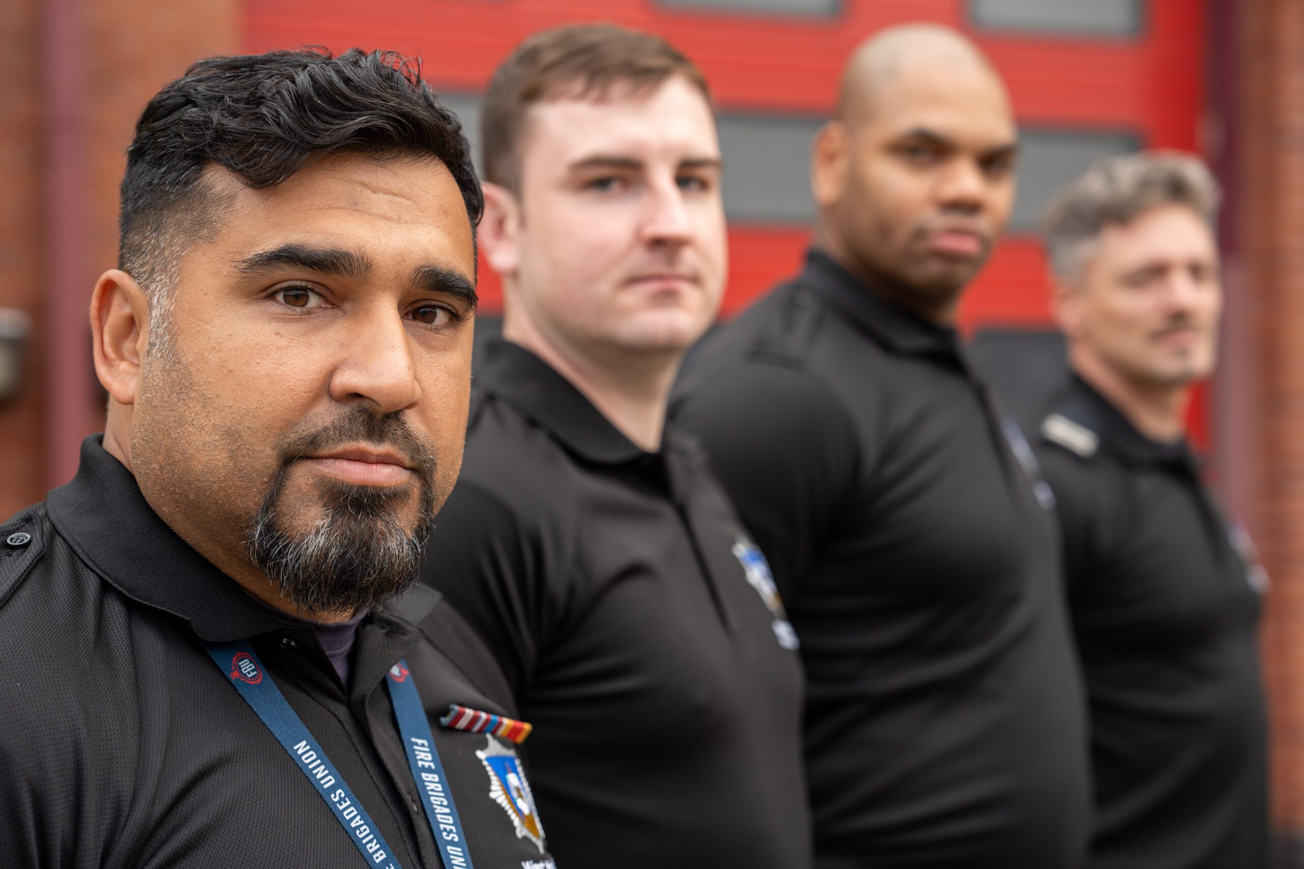 Four men in black uniforms stand in a row outdoors, facing the camera. The focus is on the man in front, while the others are slightly out of focus. A red door and brick wall are in the background.