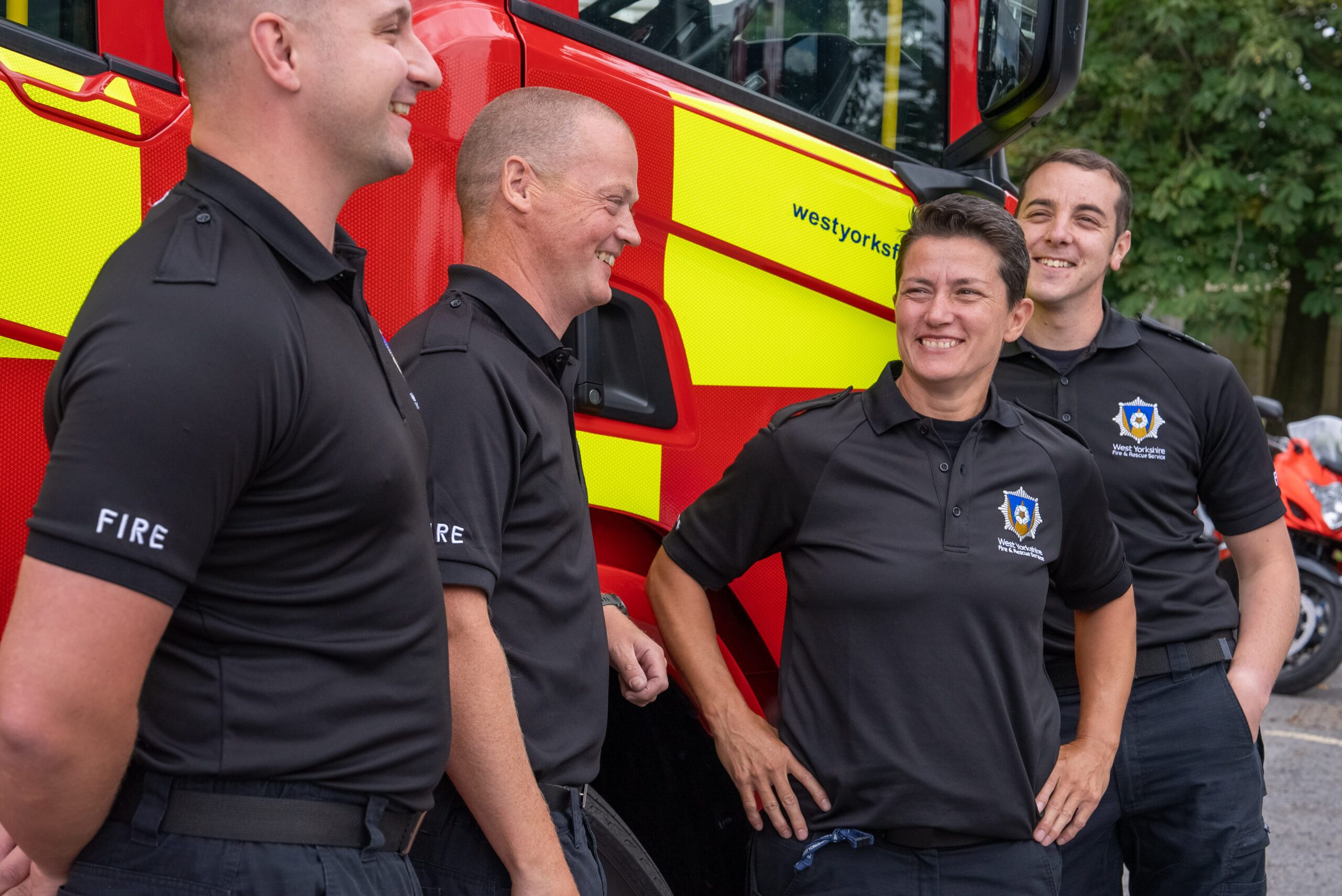 Four firefighters in uniform stand and smile together beside a red and yellow fire truck, enjoying a light moment outdoors. Trees are visible in the background. 