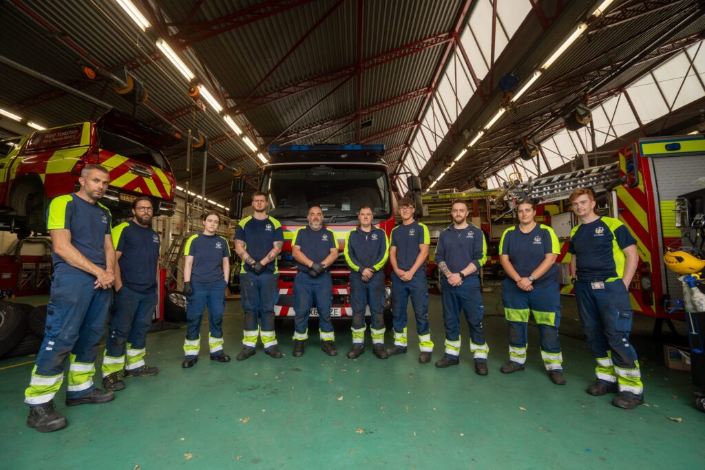 Ten firefighters in uniform stand in a row inside a fire station, with fire trucks and equipment visible in the background under a high, metal roof.