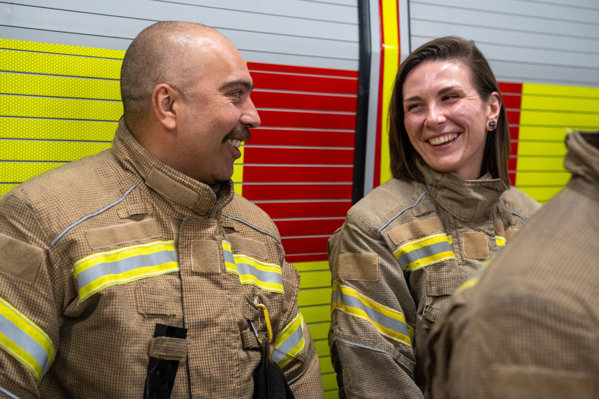 Two firefighters in brown uniforms with reflective stripes smile and talk, standing in front of a yellow and red fire truck.