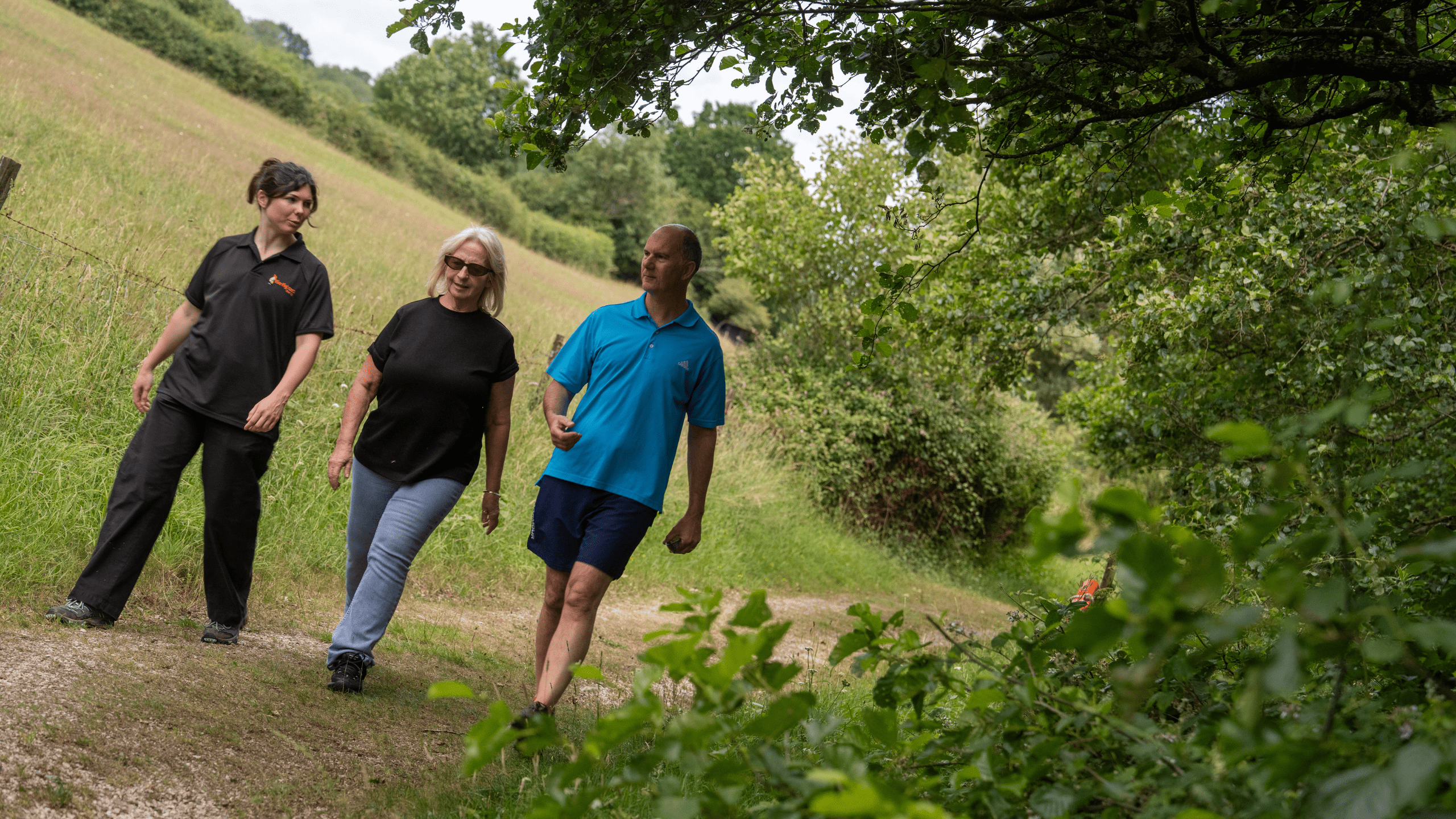 Three adults, two women and one man, walk together on a grassy forest path, surrounded by trees and greenery on a sunny day.
