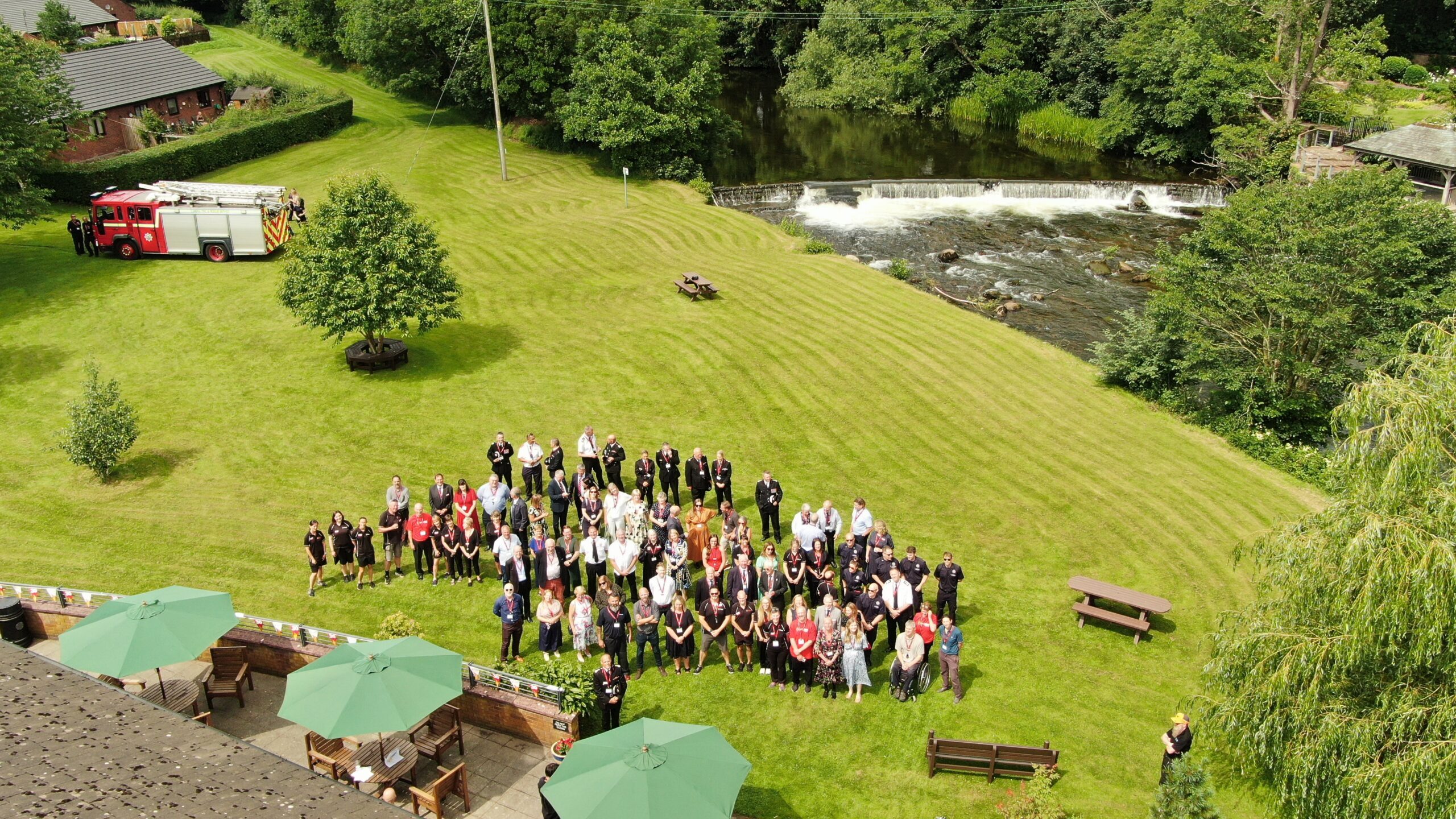 A large group of people stands on a grassy lawn near a river and waterfall, with a fire truck parked nearby and several green umbrellas shading tables in the foreground.