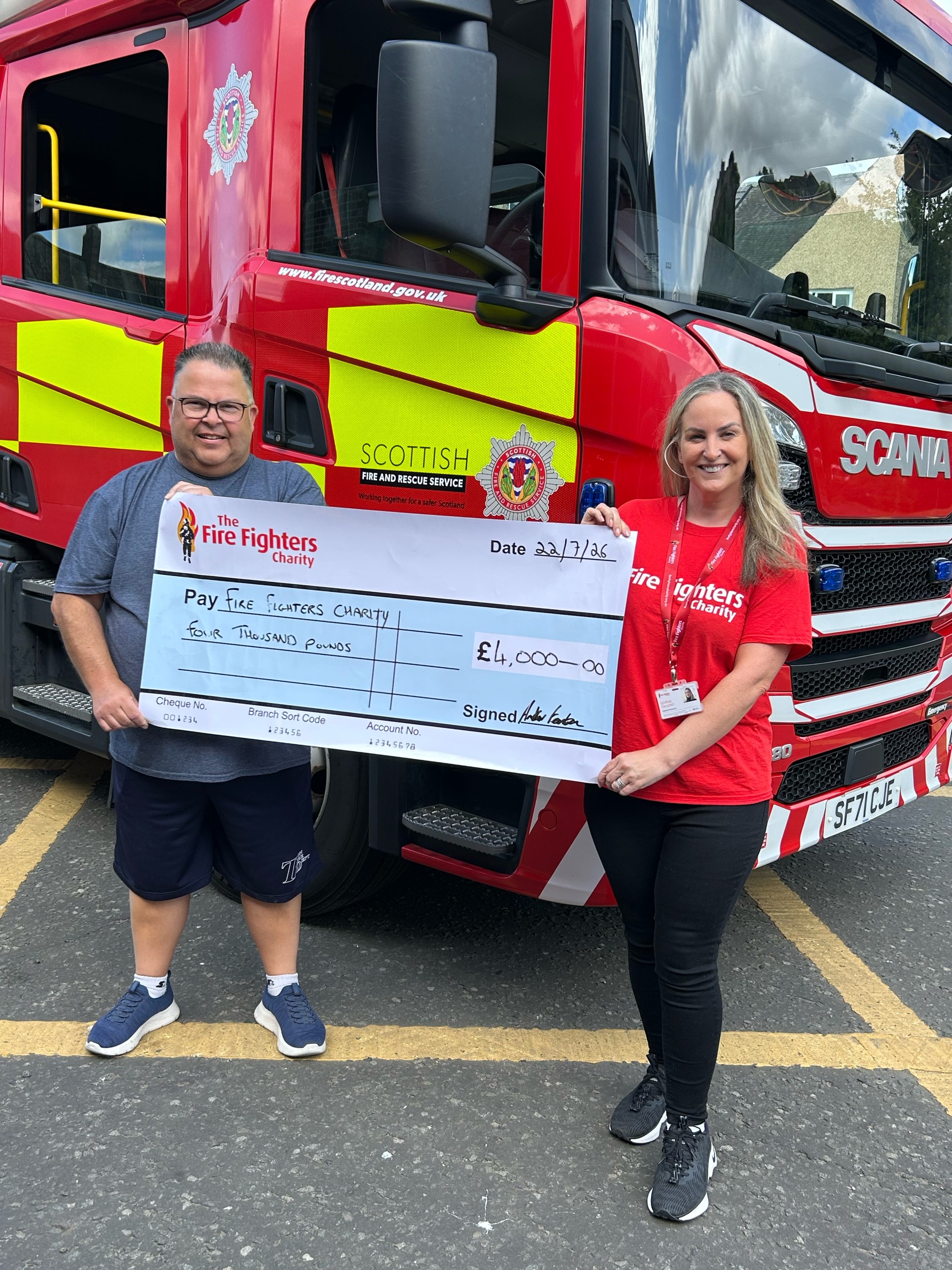 Two people stand in front of a fire engine holding a large check for £4,000 made out to The Fire Fighters Charity, dated 22/07/24. Both are smiling and wearing red charity shirts.