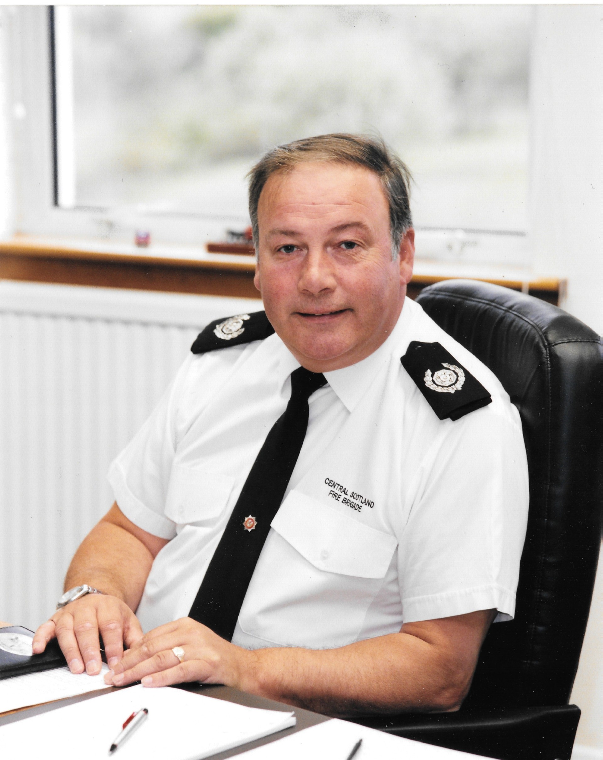 A man in a police uniform sits at a desk, looking at the camera with a slight smile. He has short hair, and the uniform reads “Central Scotland Fire Brigade.” Papers and a pen are visible on the desk.
