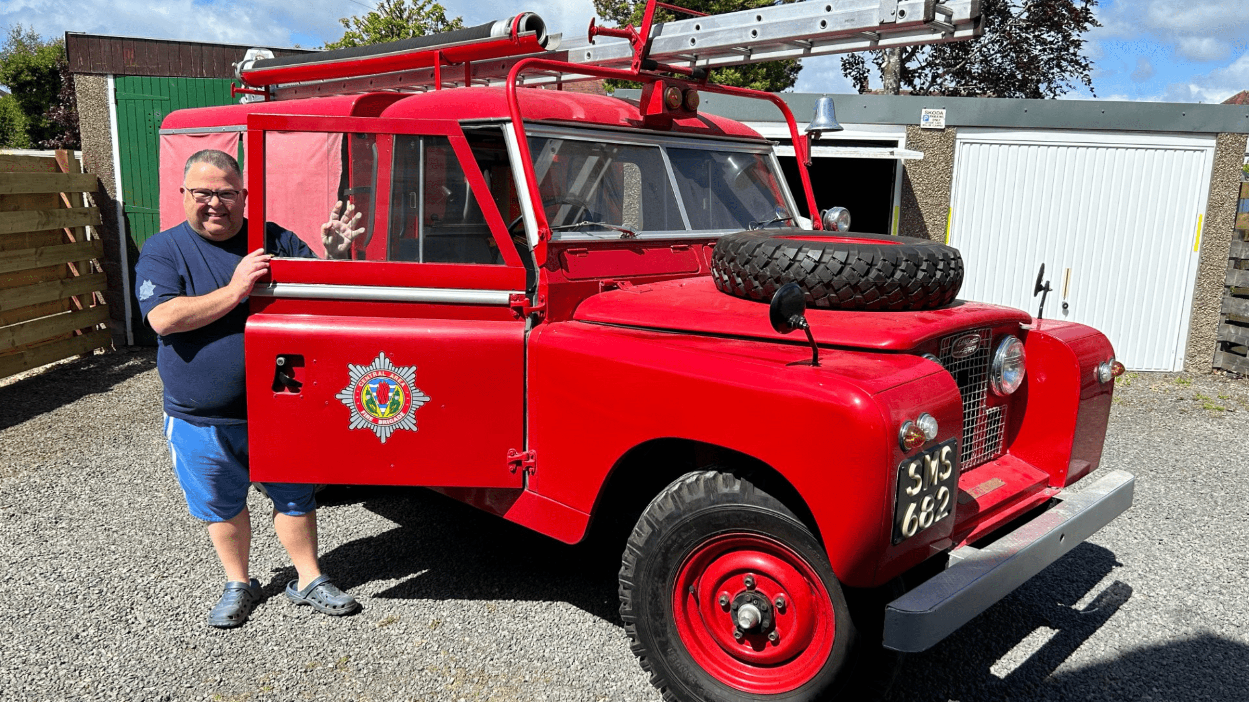 A man stands next to a vintage red fire truck with a ladder on top and a tire on the hood, smiling and waving. The truck is parked on gravel near garages.