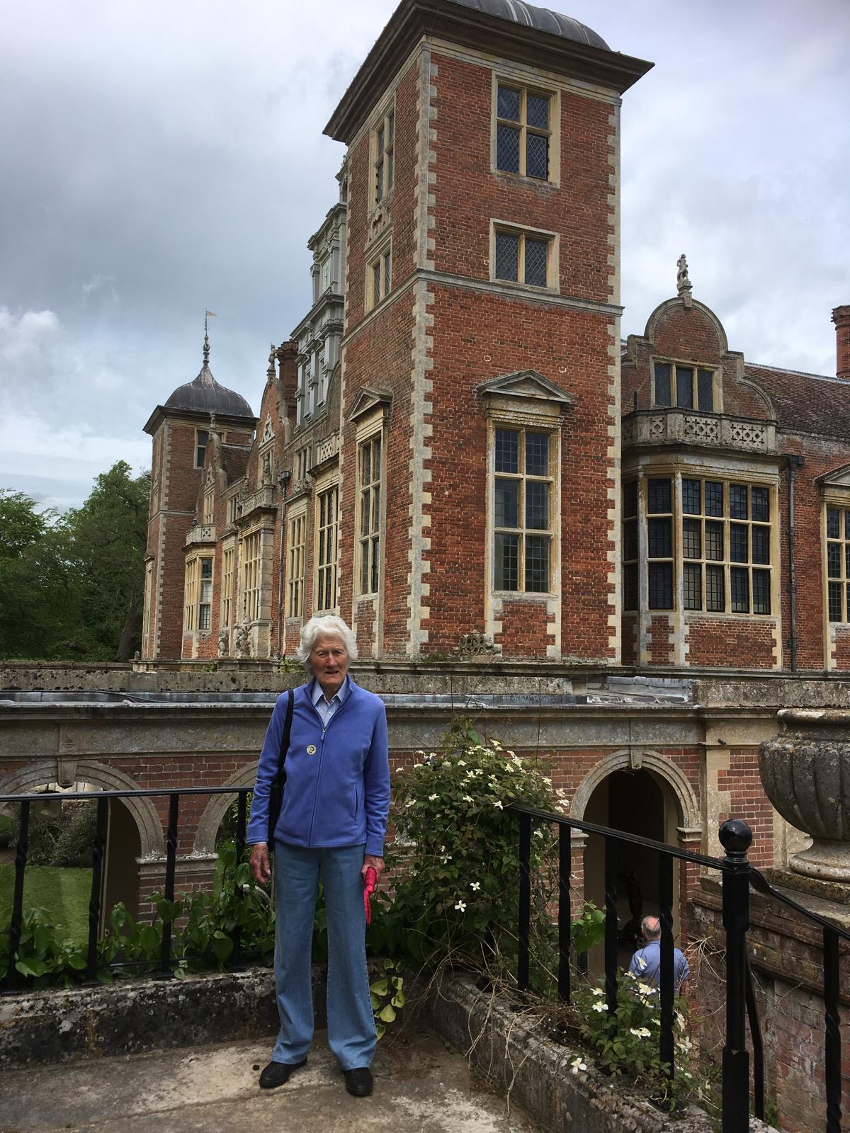 An older woman in a blue jacket and jeans stands in front of a large, ornate brick building with tall windows and towers, under a cloudy sky.