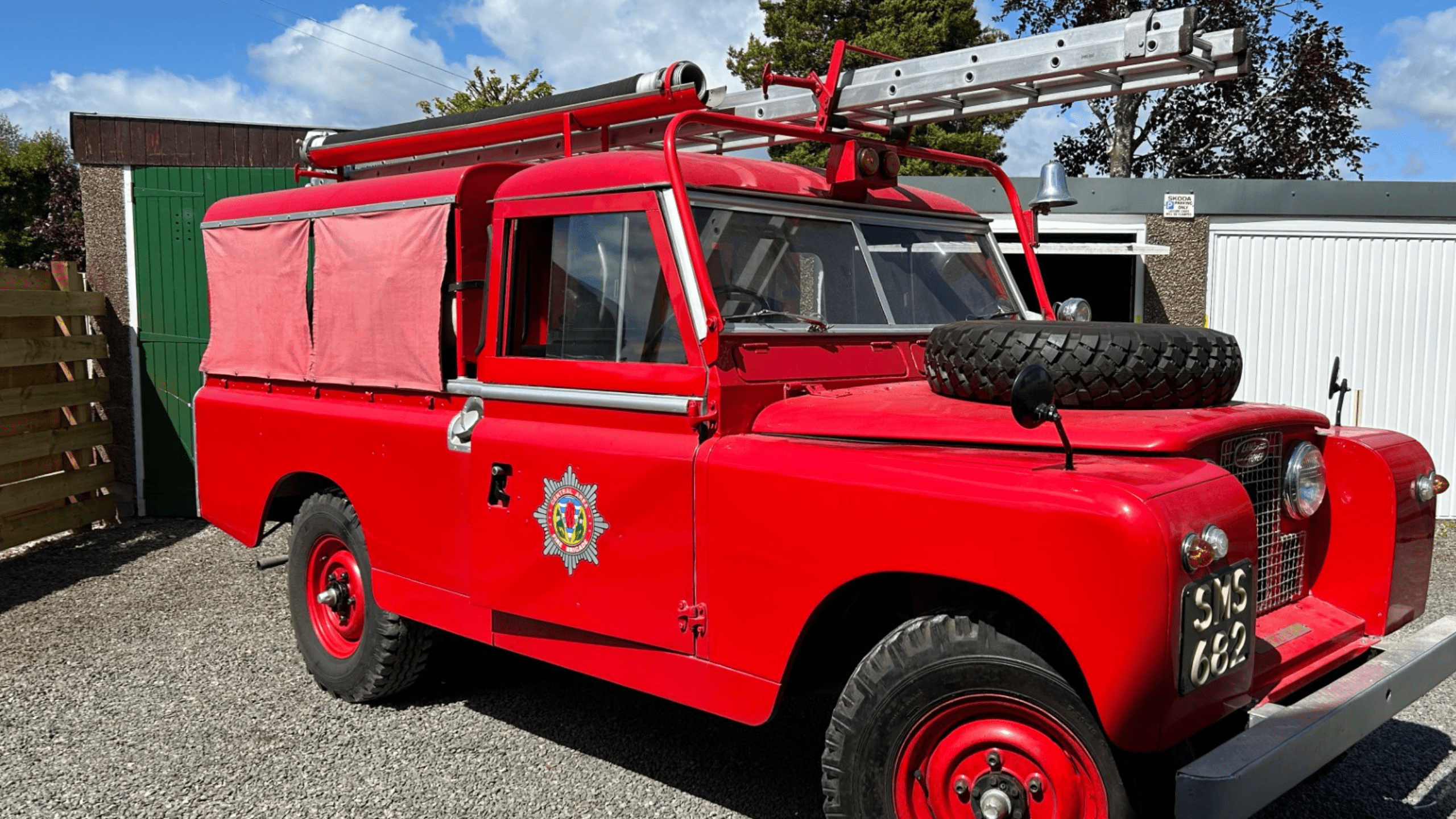 A vintage red fire engine with a ladder on top and a spare tire on the hood is parked on gravel in front of garages.