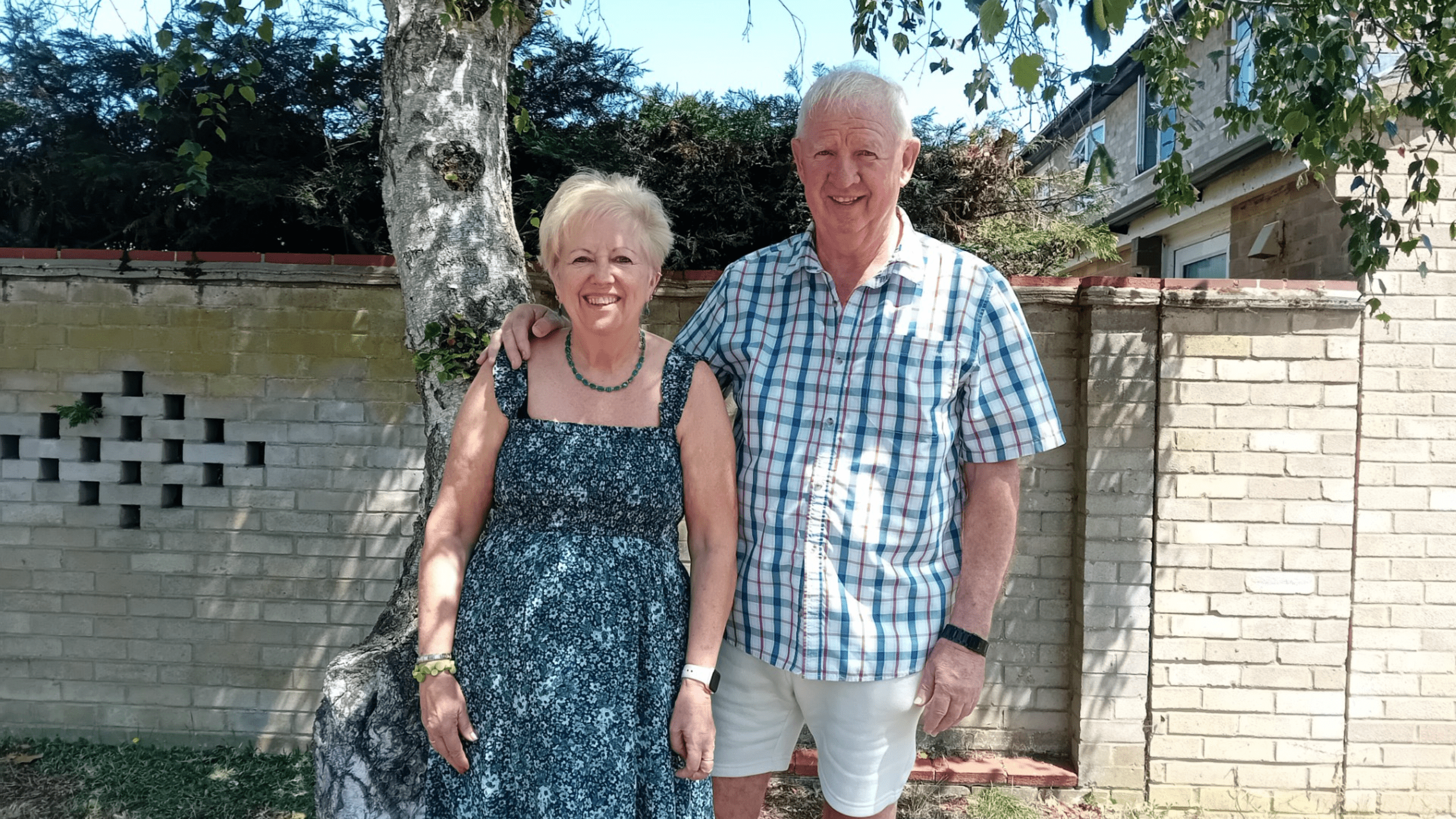 An older woman in a floral dress and an older man in a plaid shirt and shorts stand smiling together outside, next to a tree and a brick wall, with greenery and a building in the background.
