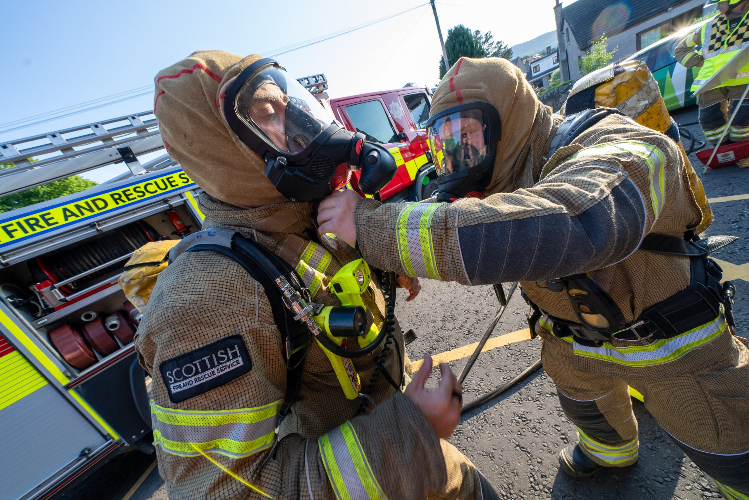 Two firefighters in full gear and breathing masks assist each other near a fire truck labeled “Scottish Fire and Rescue Service,” with equipment and vehicles in the background.